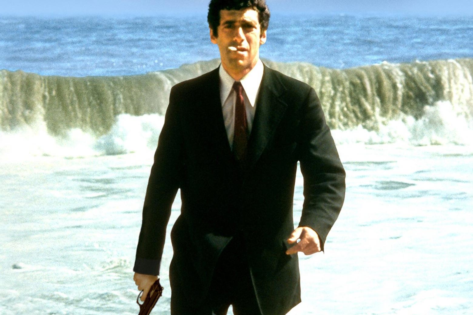 Elliot Gould walks in a suit on a beach, holding a gun